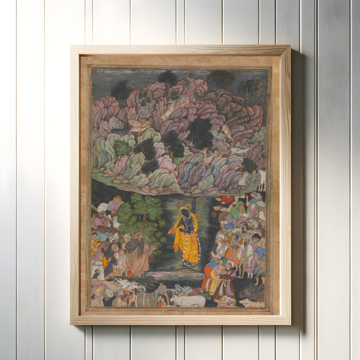 Framed art print of Krishna Holds Up Mount Govardhan to Shelter the Villagers of Braj, in a light wood frame on a white panelled wall