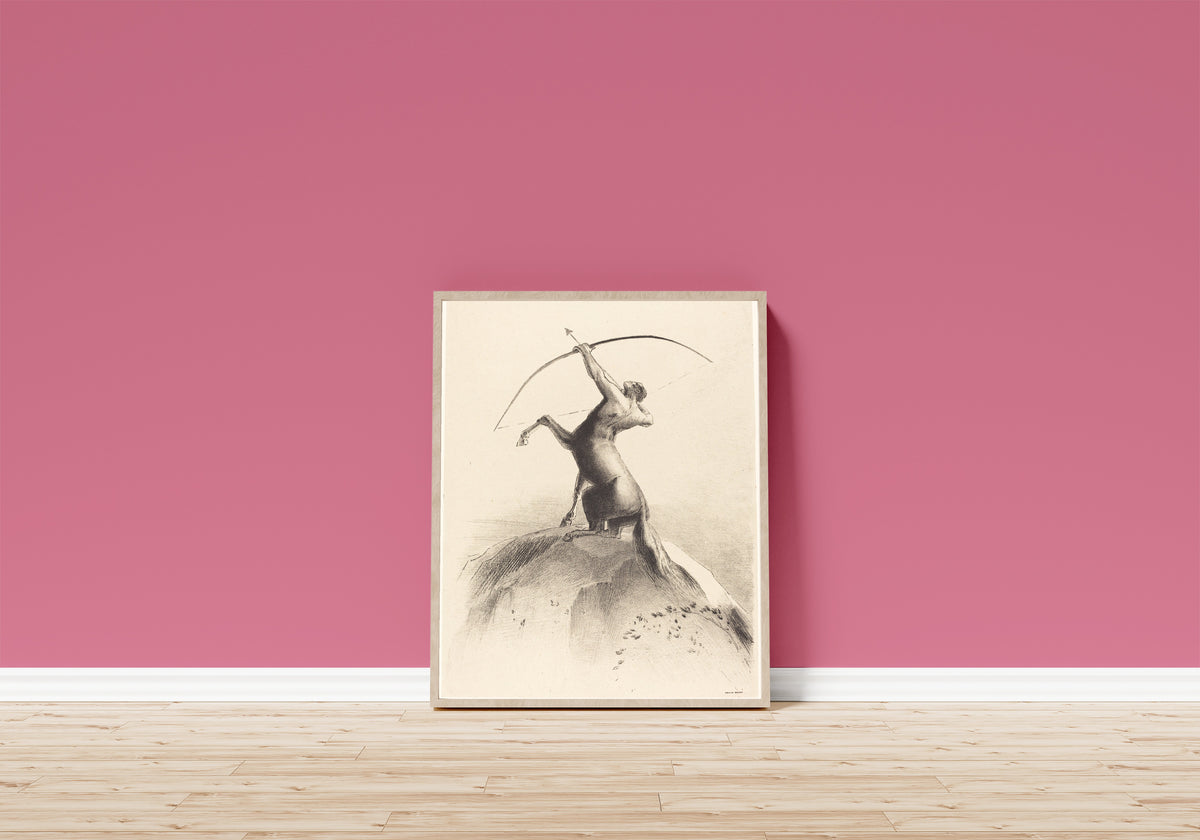 Framed art print of Centaur Aiming at the Clouds by Odilon Redon, in a light wood frame leaning on a pink wall above a wooden floor