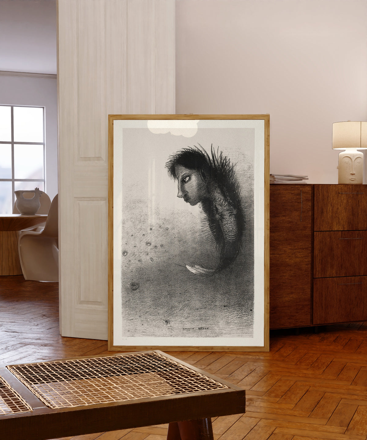 Framed art print of The Temptation of Saint Anthony: Then There Appears a… by Odilon Redon, in a light wood frame leaning in a warm room with a parquet floor