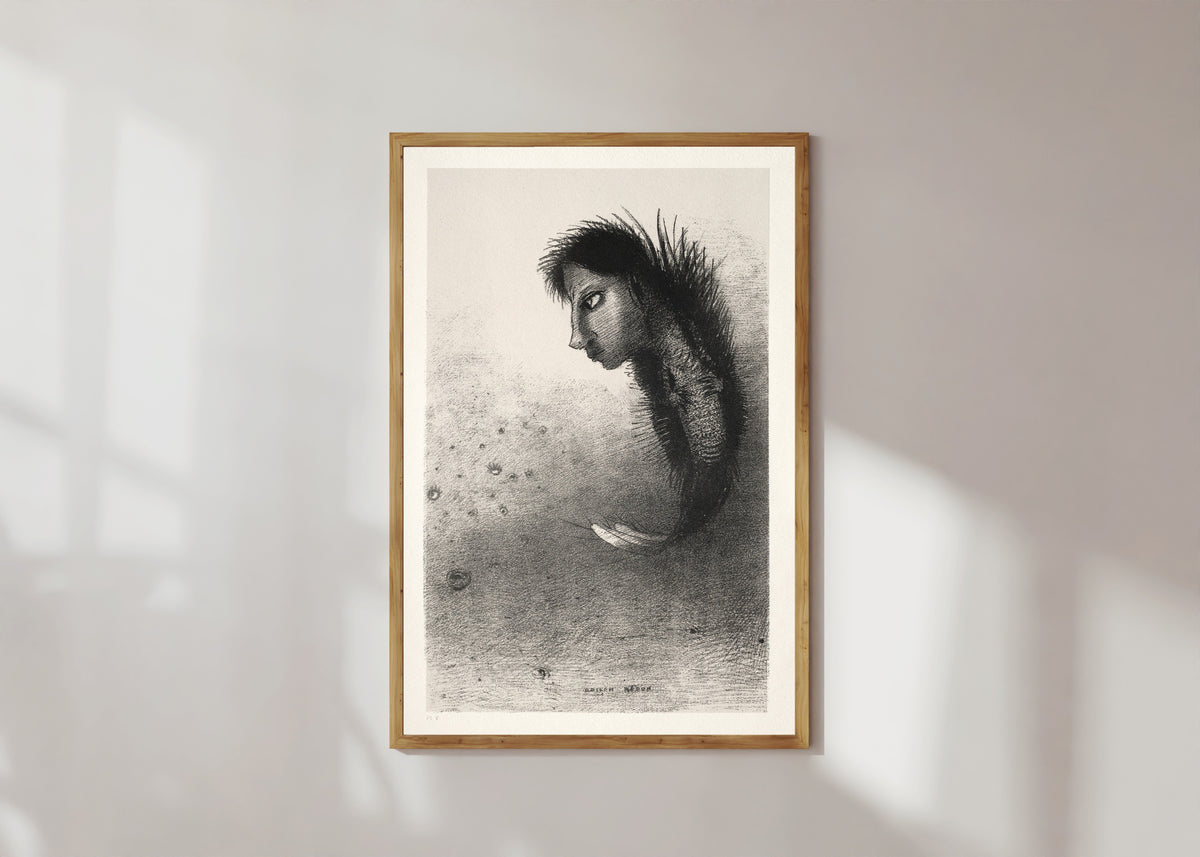 Framed art print of The Temptation of Saint Anthony: Then There Appears a Singular Being by Odilon Redon, in a light wood frame on a sunlit white wall