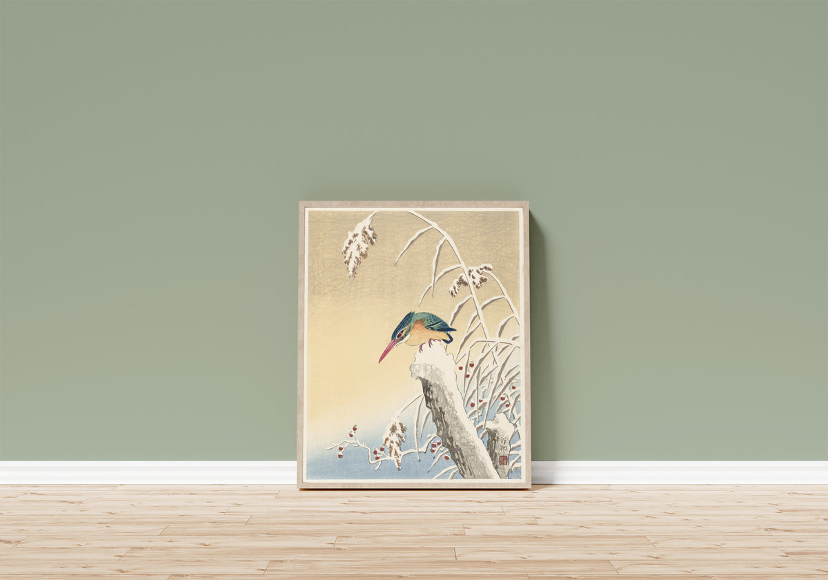 Framed art print of Kingfisher in the Snow by Ohara Koson, in a light wood frame leaning on a sage green wall above a wooden floor