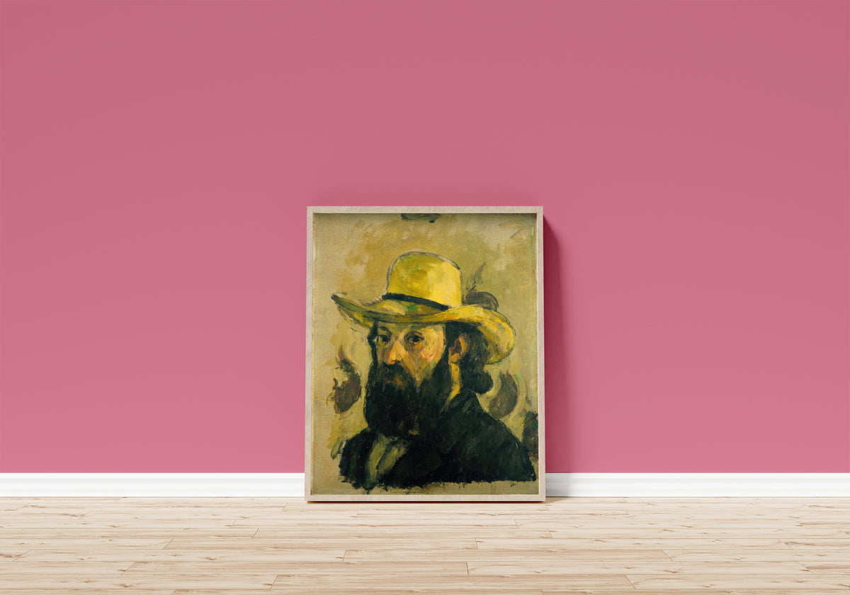 Framed art print of Self-Portrait in a Straw Hat by Paul Cezanne, in a light wood frame leaning on a pink wall above a wooden floor