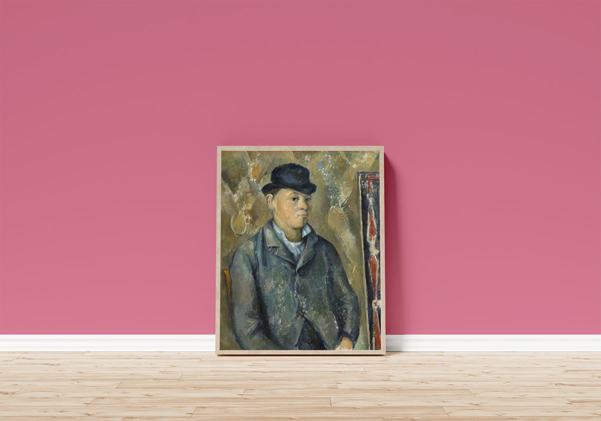 Framed art print of The Artist's Son, Paul by Paul Cezanne, in a light wood frame leaning on a pink wall above a wooden floor