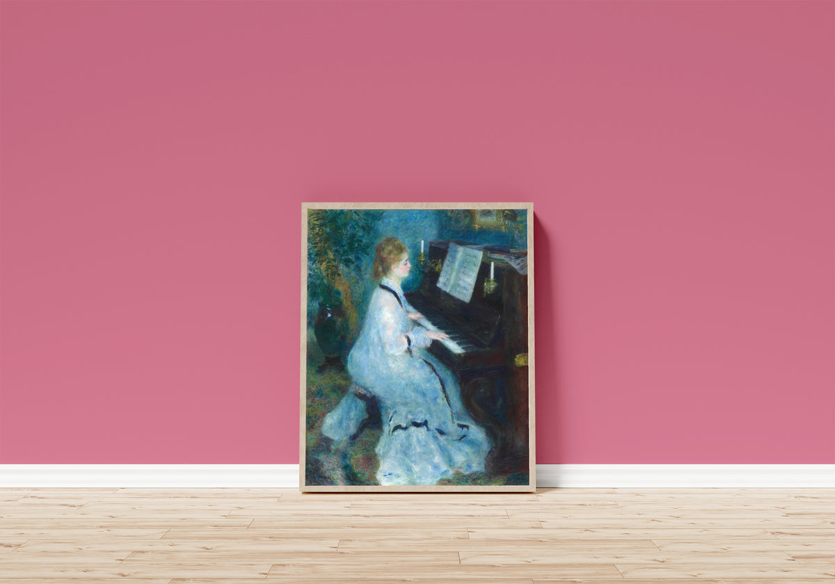 Framed art print of Woman at the Piano by Pierre-Auguste Renoir, in a light wood frame leaning on a pink wall above a wooden floor