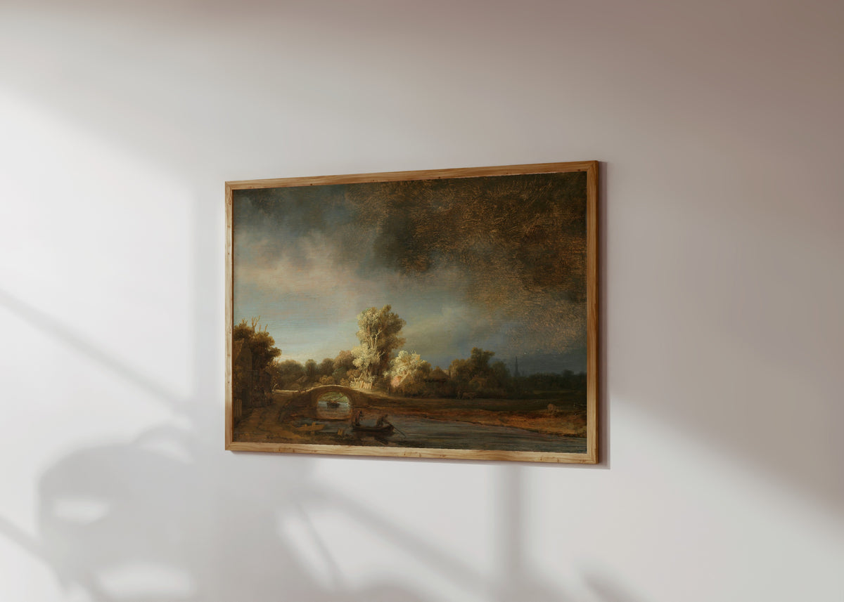 Framed art print of Landscape with a Stone Bridge by Rembrandt van Rijn, in a light wood frame on a sunlit white wall, seen at a slight angle