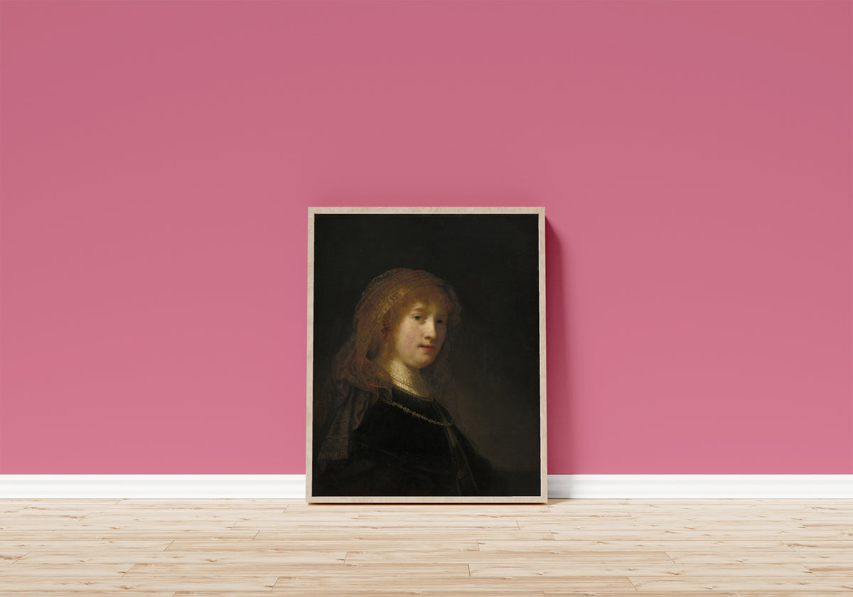 Framed art print of Saskia van Uylenburgh, the Wife of the Artist by Rembrandt van Rijn, in a light wood frame leaning on a pink wall above a wooden floor