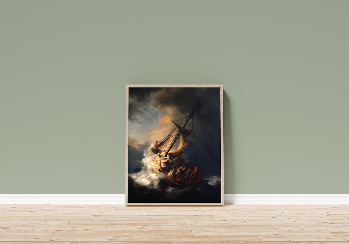 Framed art print of The Storm on the Sea of Galilee by Rembrandt van Rijn, in a light wood frame leaning on a sage green wall above a wooden floor