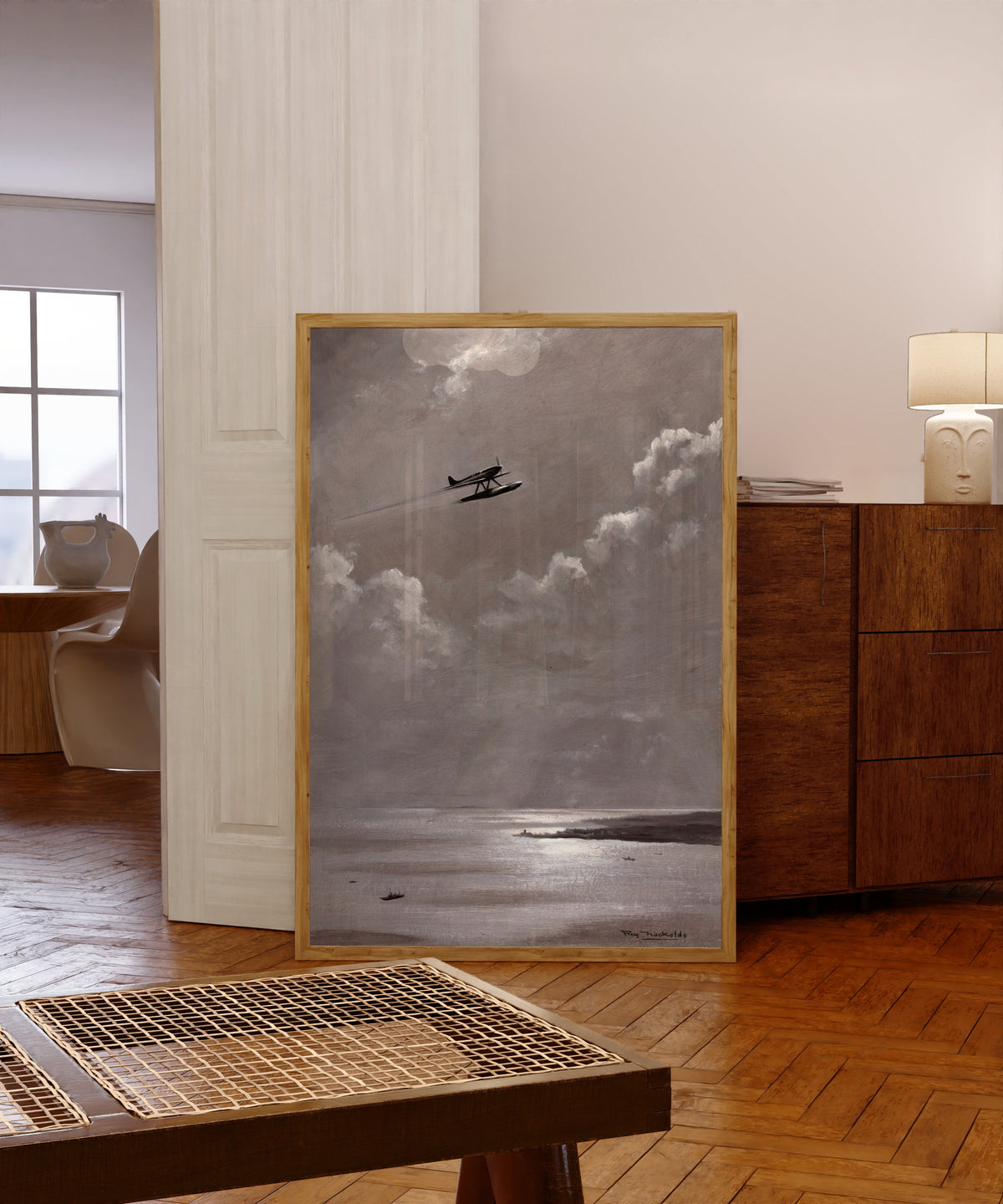 Framed art print of Seaplane over the Coast by Roy Nockolds, in a light wood frame leaning in a warm room with a parquet floor