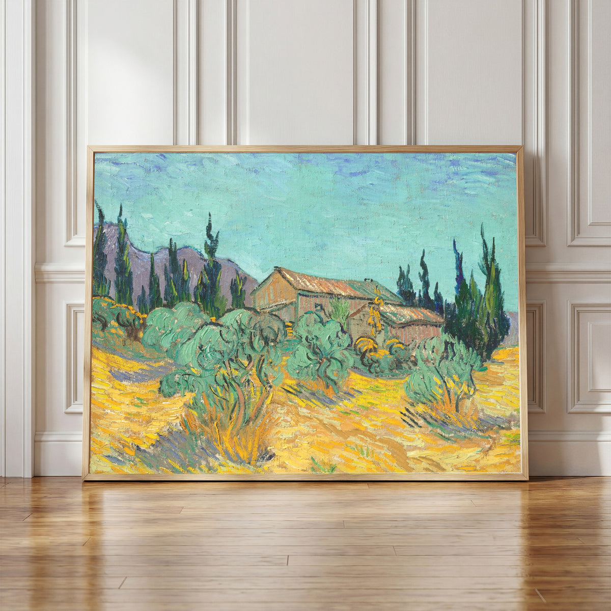 Framed art print of Huts surrounded by Olive Trees and Cypresses by Vincent van Gogh, in a light wood frame leaning against a panelled white wall