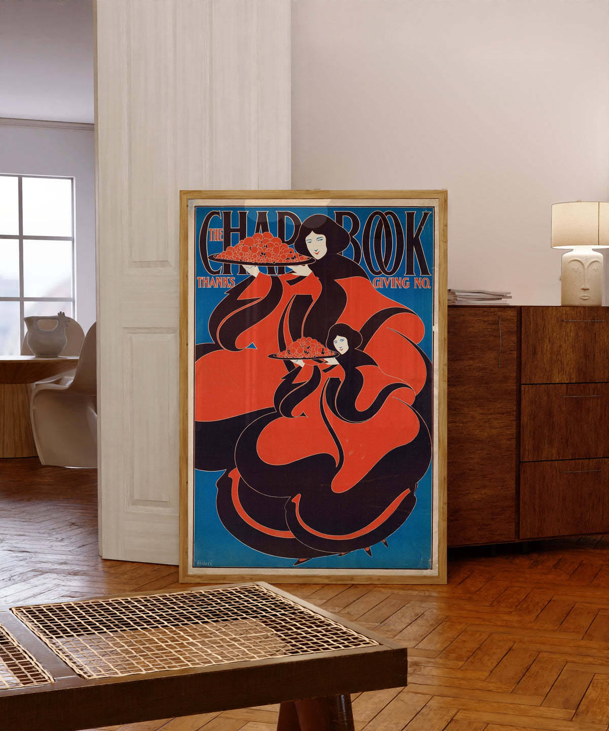 Framed art print of The Chap-Book, Thanksgiving Number, 1895 by Will Bradley, in a light wood frame leaning in a warm room with a parquet floor